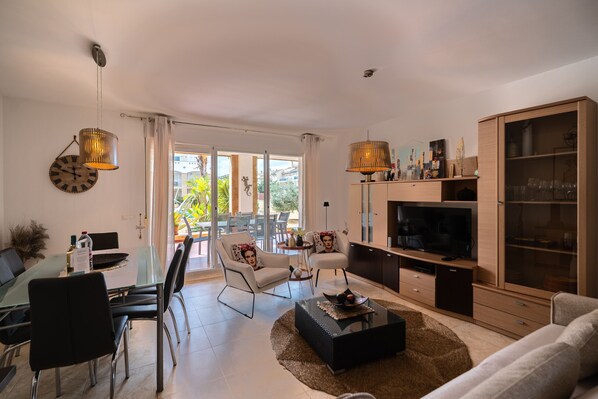 A modern living room with a glass dining table, two white armchairs, a black coffee table, a TV, and a wooden cabinet. - Pompidú Apartment (Javea)