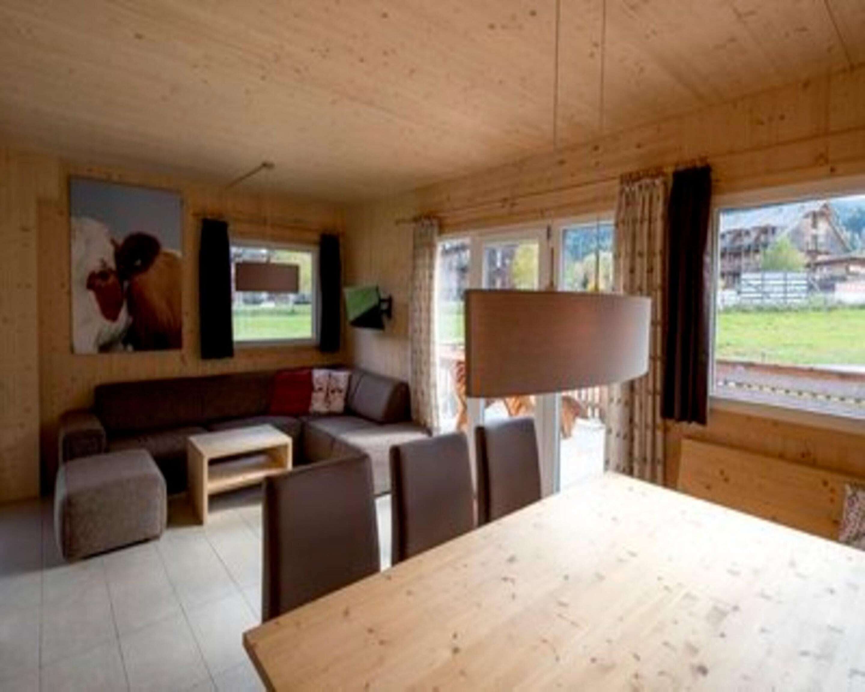 A cozy cabin interior with a wooden dining table, a sofa, and a large window offering a view of the outdoors.