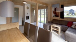 A modern, open-plan living and dining area with wooden walls and ceiling, a large window with a view of the outdoors, a fireplace, and a mounted television.