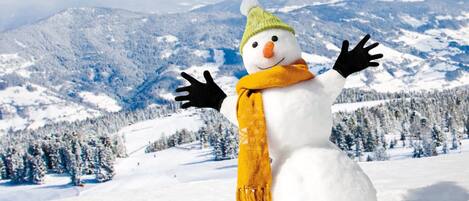 A snowman with a green hat and yellow scarf in a snowy landscape with mountains in the background.