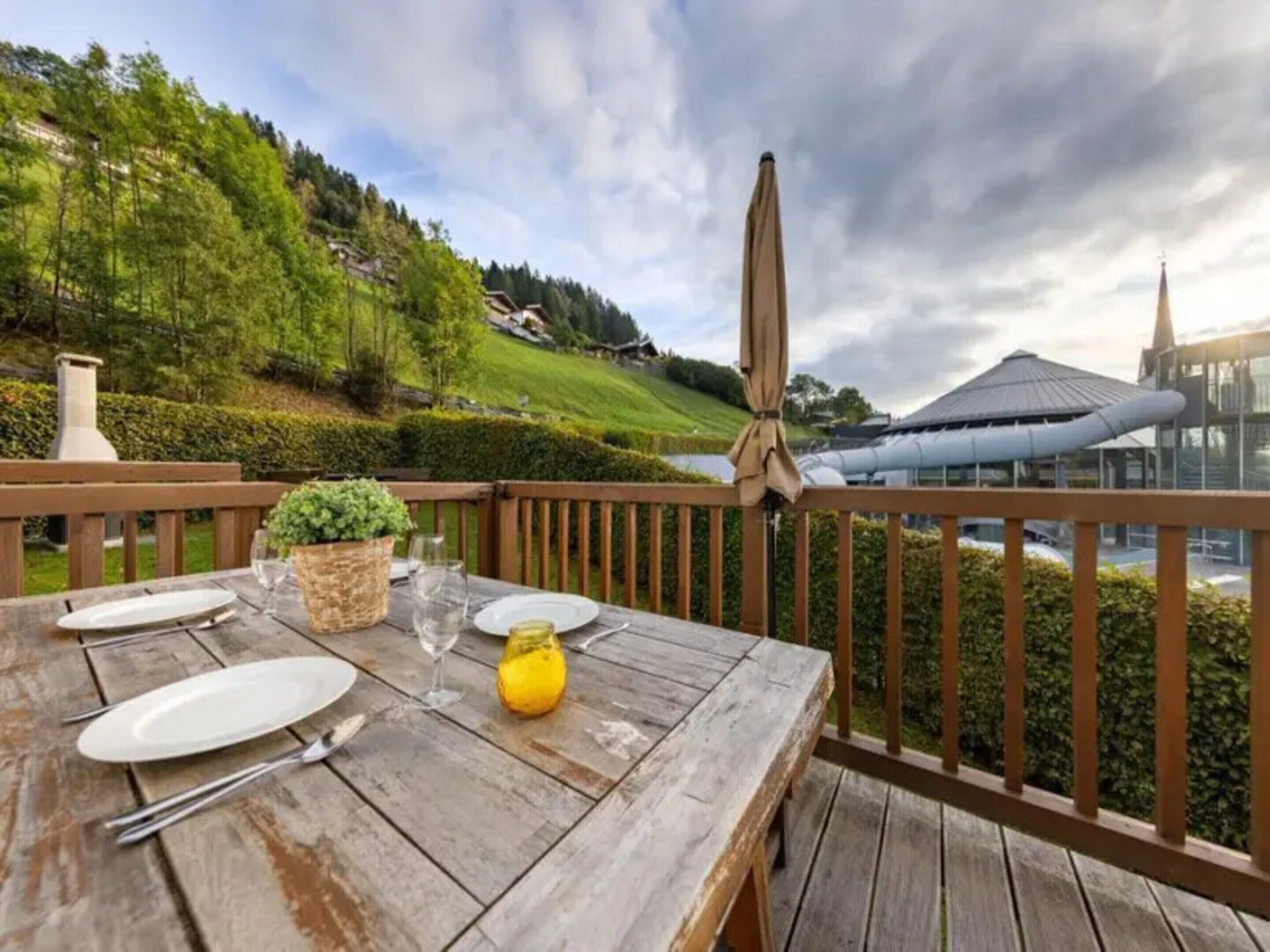 Eine Holzterrasse mit Esstisch, mit Blick auf eine grüne Landschaft und ein modernes Gebäude mit einem Turm.