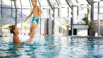 Ein überdachter Schwimmbereich mit großen Glaswänden, Blick ins Freie, und eine Person steht auf den Schultern einer anderen.