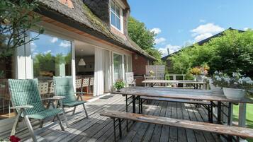 Une terrasse en bois avec des meubles d’extérieur, un bâtiment en briques surmonté d’un toit de chaume et un espace jardin agrémenté de plantes en pot.