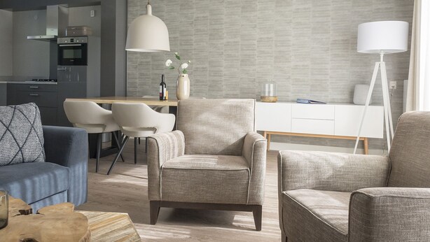 A modern living room with a dining area, a kitchen in the background, and a large window providing natural light.