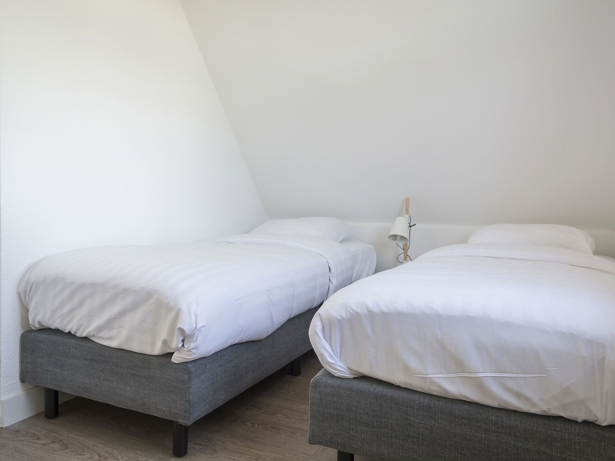 Ein Zimmer mit zwei Einzelbetten, jeweils mit weißer Bettwäsche und grauen Kopfteilen, vor einer schlichten weißen Wand.