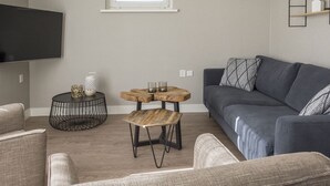 A living room with a grey sofa, a wooden coffee table, a round black side table, a TV mounted on the wall, and a window with blinds.