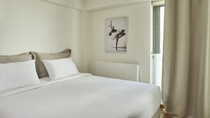 Una cama bien hecha con ropa blanca y un cabecero, un cuadro en la pared y una puerta con una cortina entreabierta.
