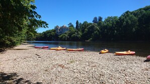 A pebbled beach with kayaks lined up, a castle-like building in the background, and dense trees surrounding the area. - Villa in Bruniquel by Aveyron River (Bruniquel)