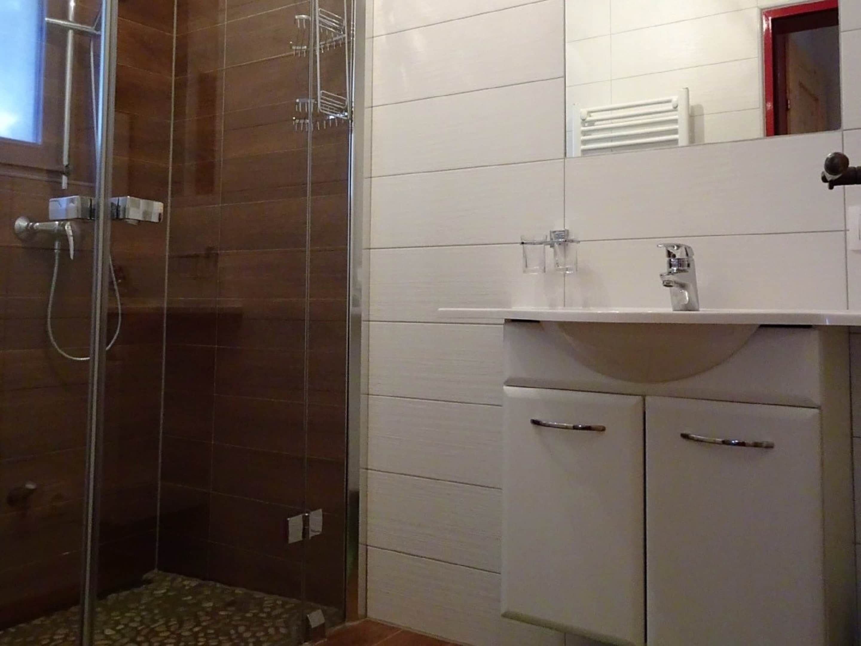 Un baño con cabina de ducha de vidrio, paredes con azulejos blancos, un lavabo blanco y un espejo sobre él.