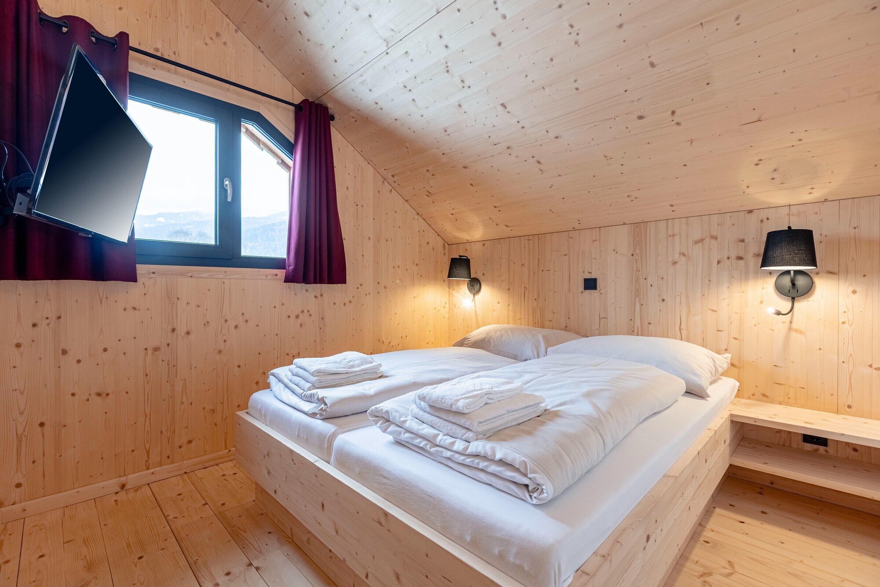 A wooden-paneled room with a bed, two wall-mounted lamps, and a window with curtains.