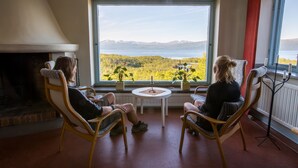 Dos sillones de mimbre mirando a una chimenea y un ventanal con vistas a un lago y montañas.