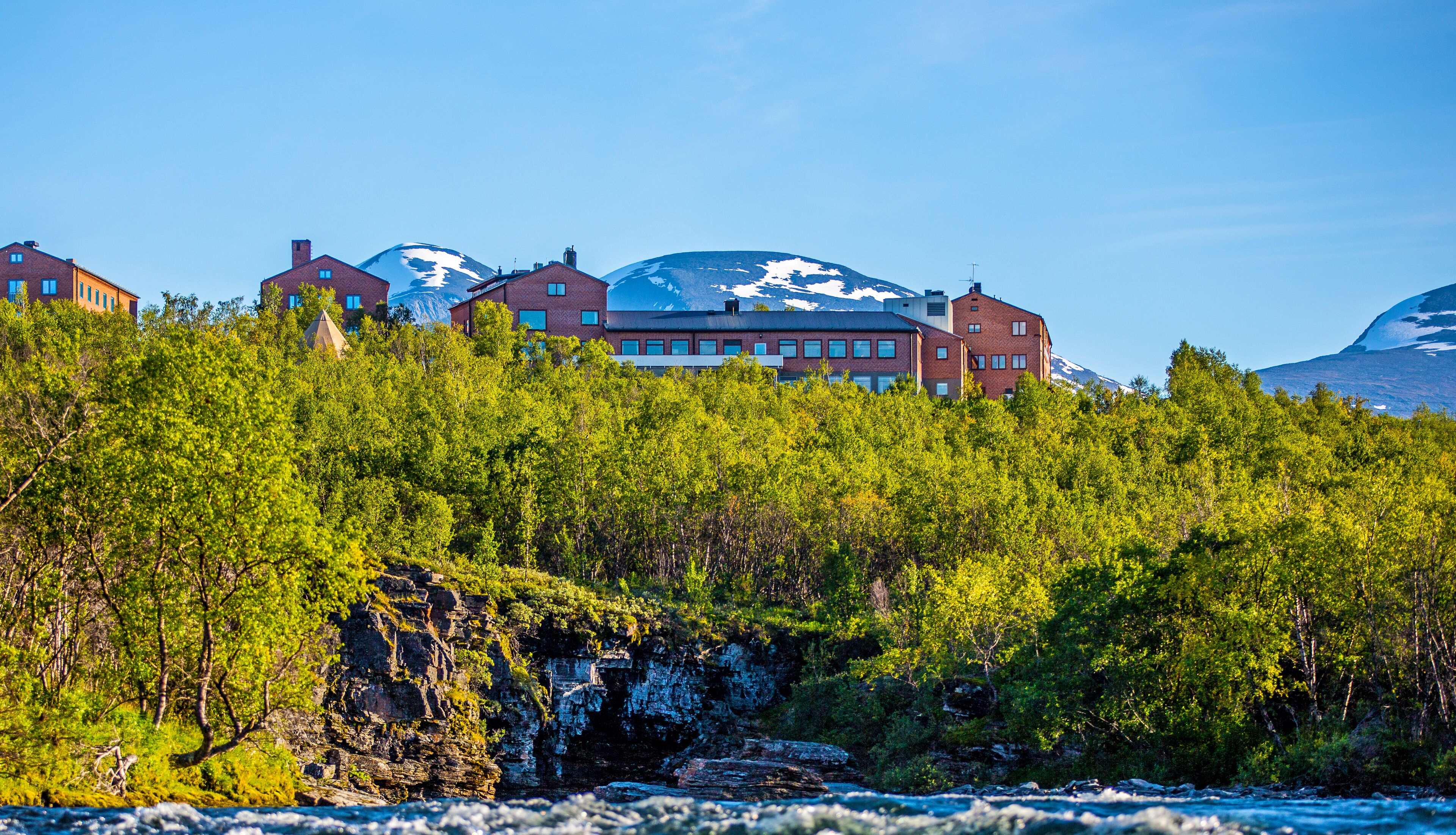 Un hotel con varios edificios de ladrillo rojo, un río en primer plano y montañas nevadas al fondo.