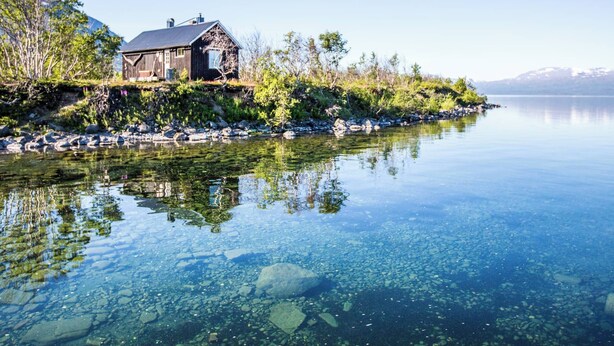 Una cabaña junto a un lago tranquilo con rocas y vegetación visibles.