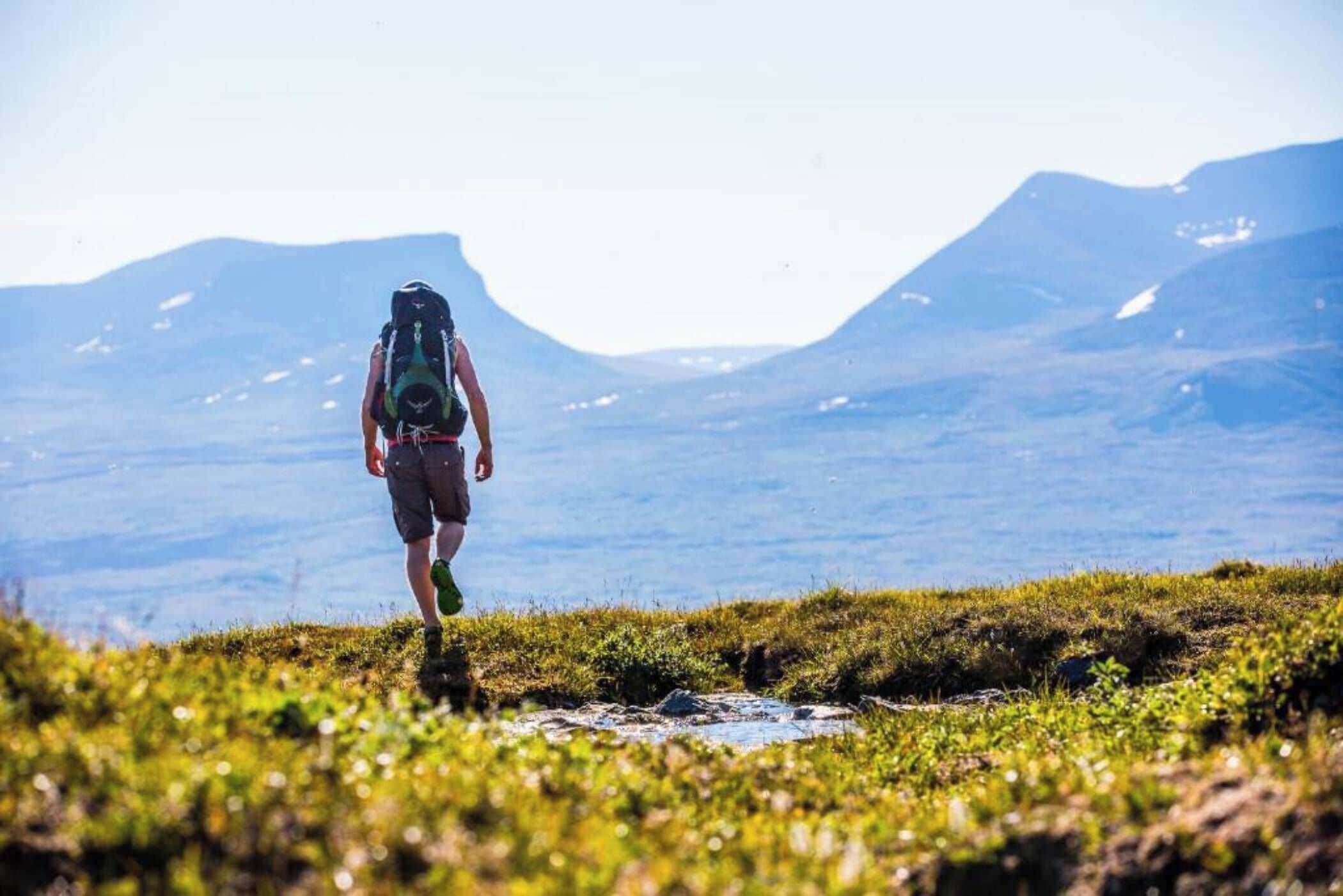 Una persona camina de excursión con una mochila sobre un terreno cubierto de hierba, con montañas al fondo.