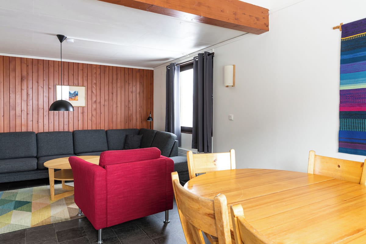 A modern living room with a wooden ceiling, a grey sofa, a red armchair, a wooden dining table, and a colorful woven tapestry.