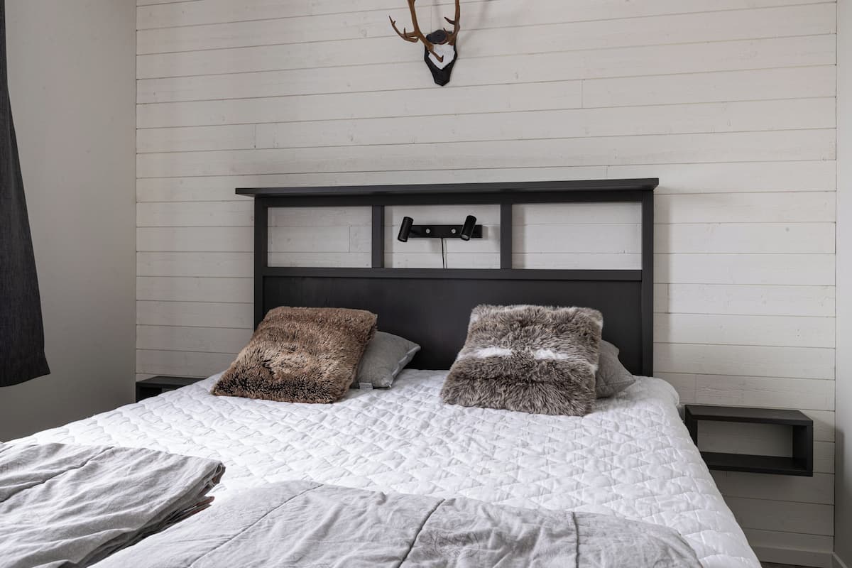 A bed with a quilted white cover, two fluffy pillows, a dark headboard, and a mounted deer head.