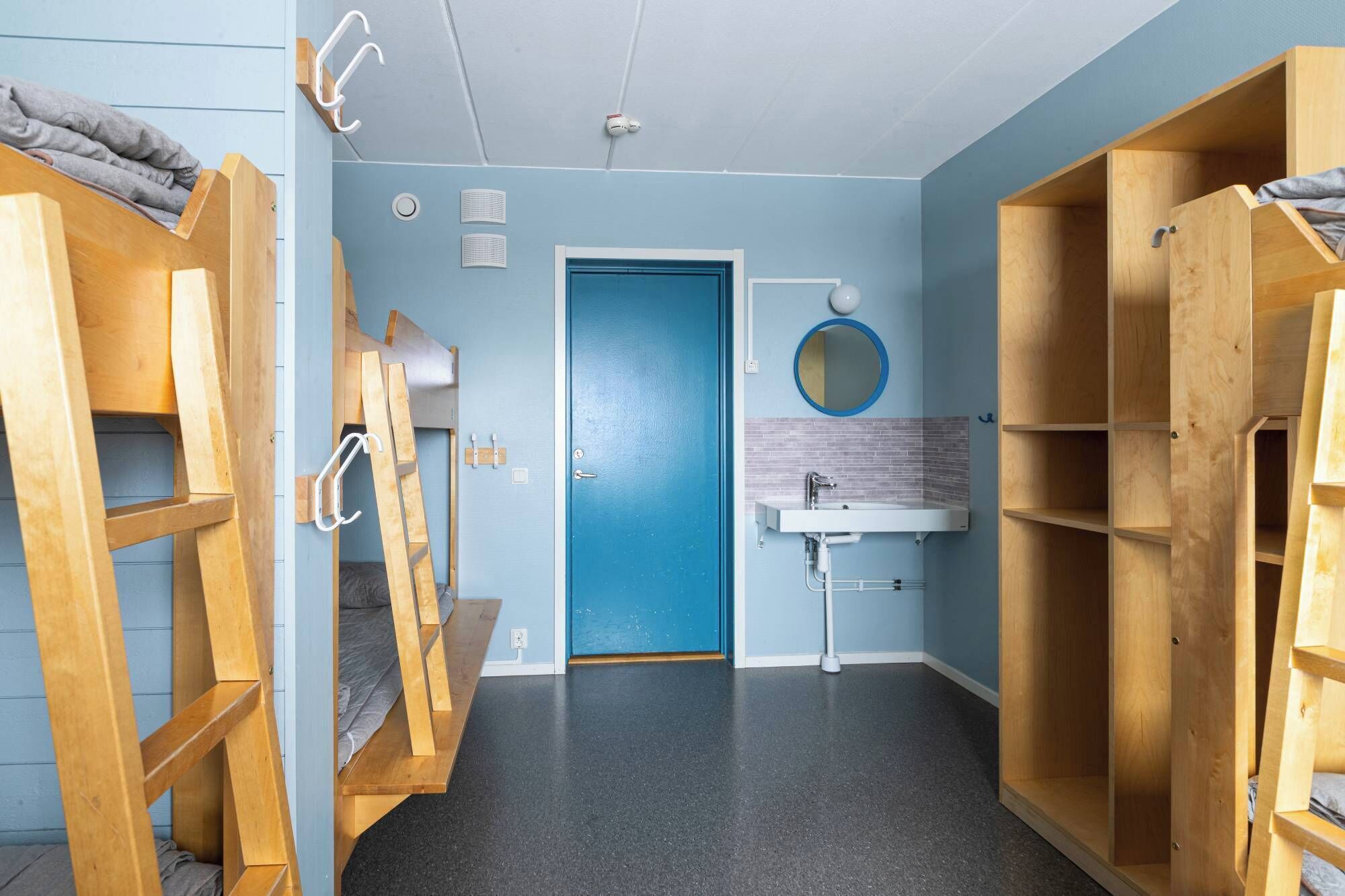 Een kleine kamer met een blauwe deur, een wastafel en een spiegel. Er zijn houten planken en een ladder.