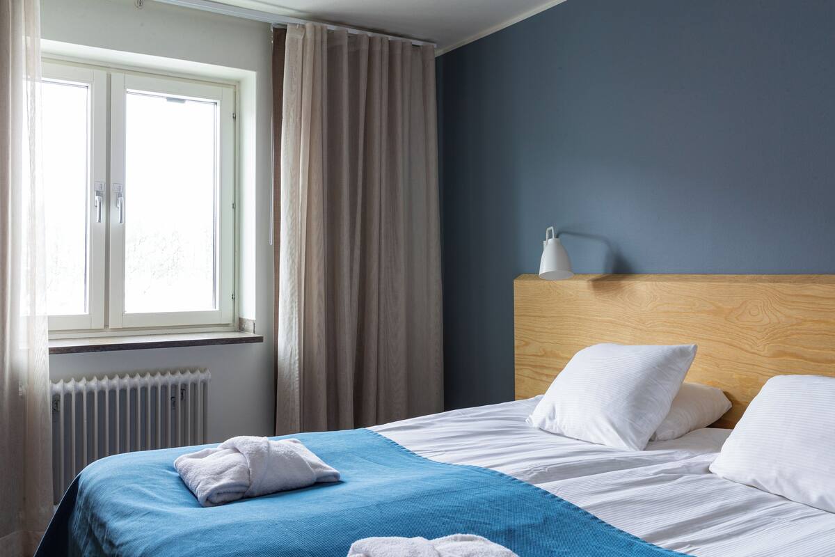 A hotel room with a bed, a window with curtains, a radiator, and a bedside table with a lamp.