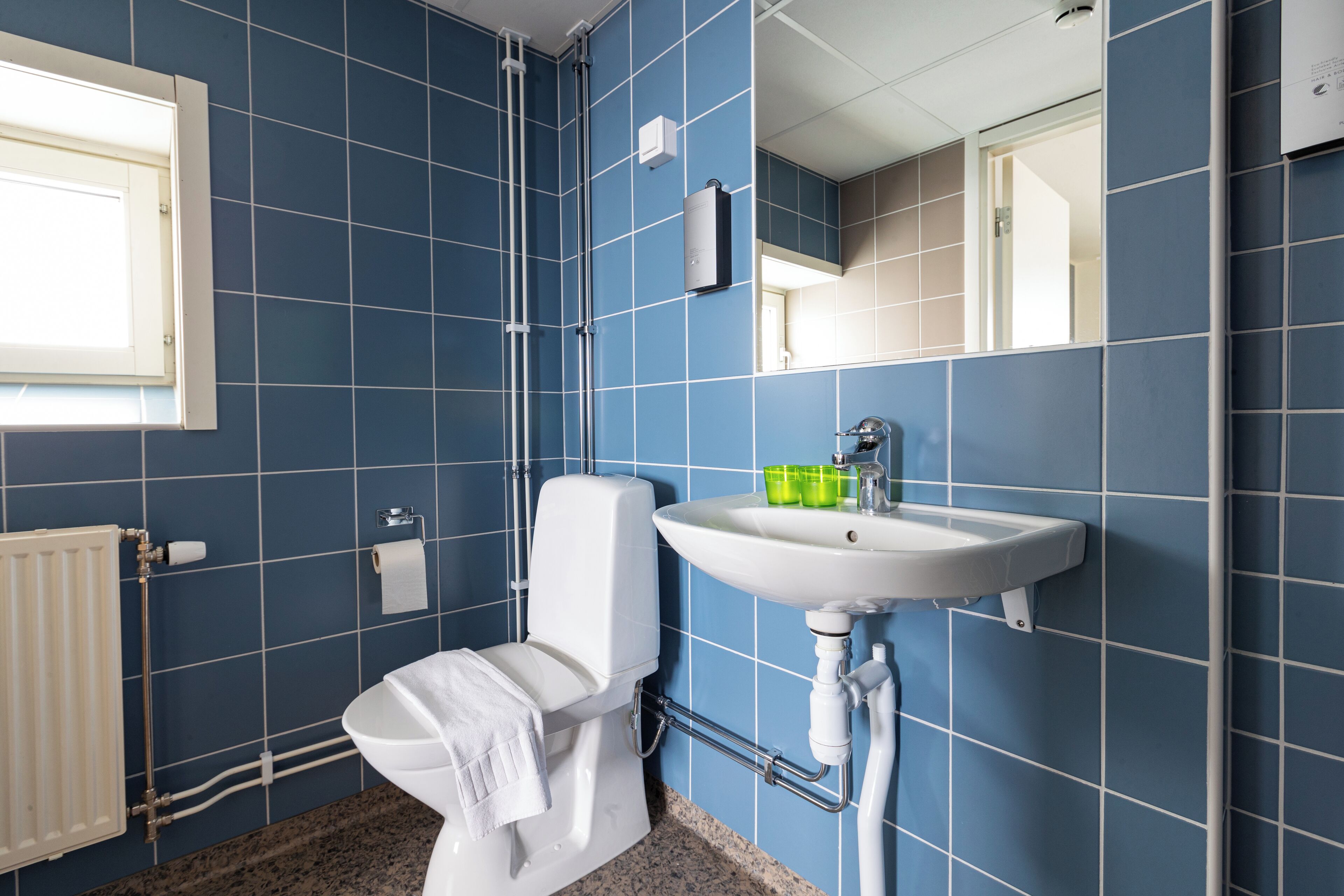 A bathroom with blue tiles, a white toilet, a sink with green cups, and a window.