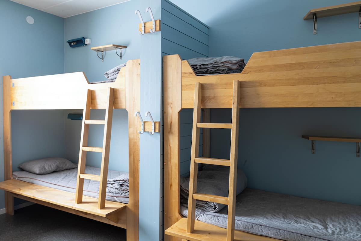 A room with two bunk beds, each with a ladder, a shelf above the beds, and a wall-mounted hook.