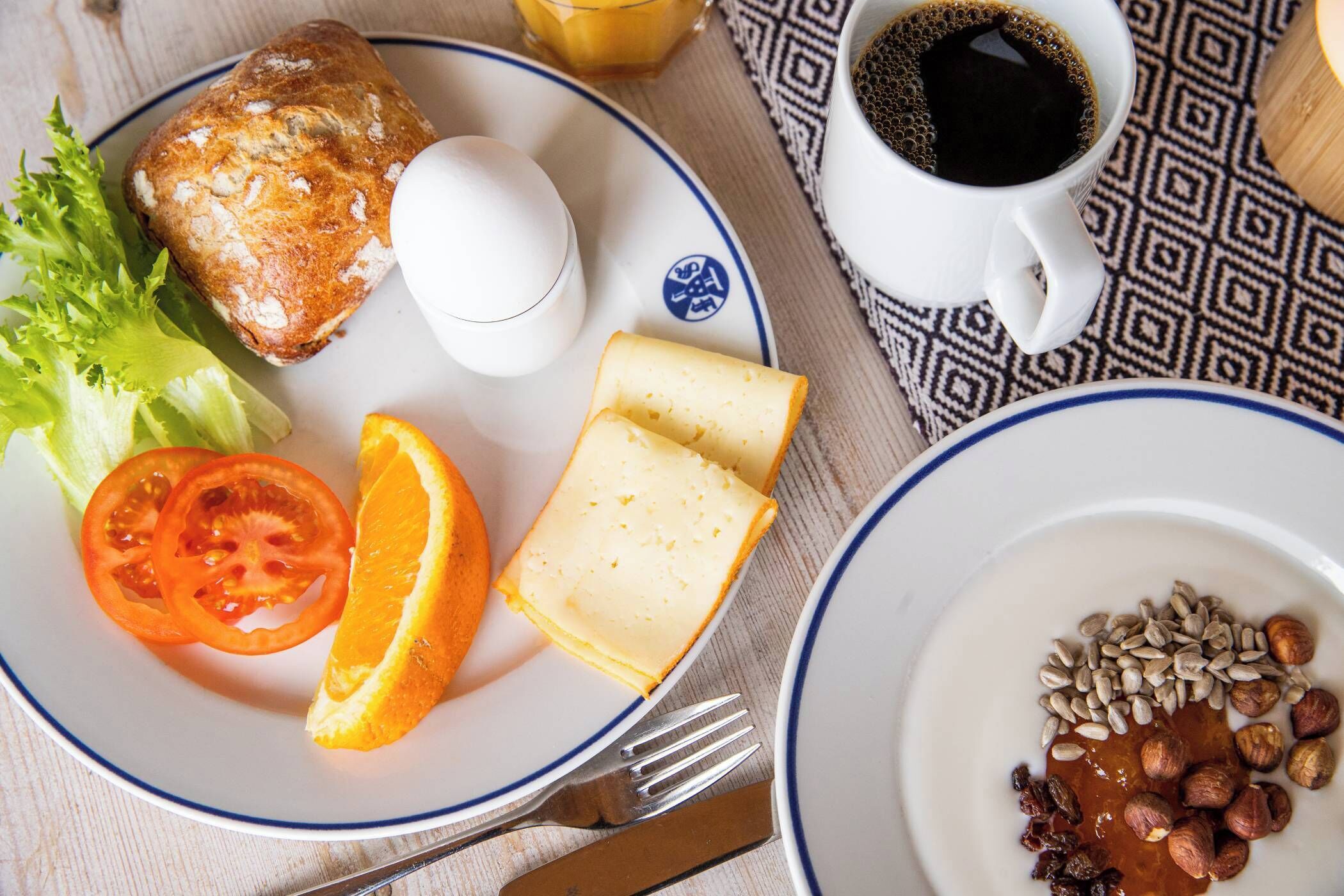 Une assiette de petit-déjeuner avec des toasts, un œuf, des tomates, une orange et du fromage, et une petite assiette d’accompagnement avec des noix et des fruits secs.