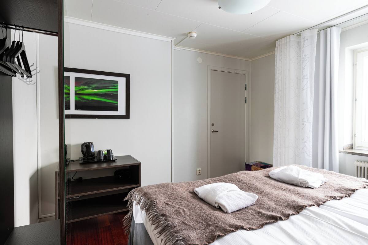 A hotel room with a bed, a shelf with items, a door, and a window with curtains.