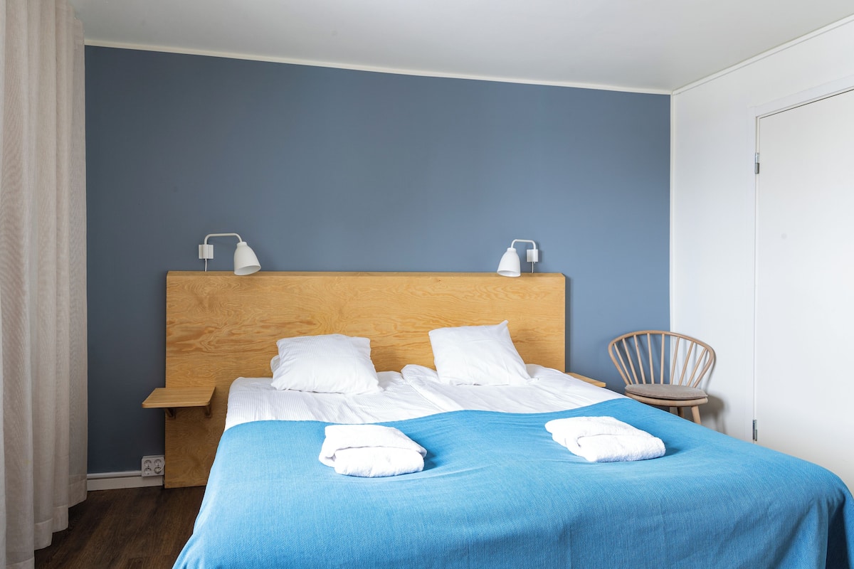 A neatly made bed with white linens and a blue bedspread, a wooden headboard, bedside tables with lamps, a wooden chair, and a white door.
