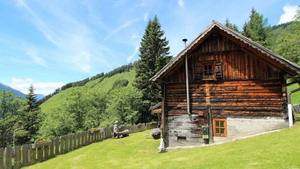 A rustic wooden cabin with a stone foundation, set on a grassy hillside with a wooden fence and a mountain backdrop. - Chalet in Obervellach in Carinthia (Obervellach)
