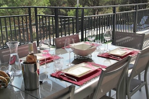 Une table à manger, dressée avec des assiettes, des verres et des couverts, entourée de chaises, sur un balcon doté d’une balustrade.