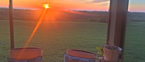 Quarto com duas cadeiras viradas para uma janela ampla, um vaso de planta e vista para um pôr do sol sobre uma paisagem gramada.
