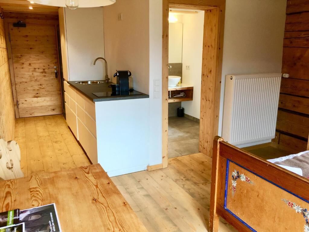 A compact room with a wooden floor, a small kitchenette with a sink and a coffee maker, a bed with a headboard, and a wooden door.