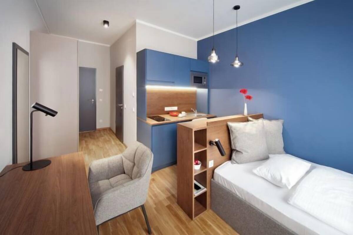 A modern hotel room with a wooden desk, a chair, a bed, a blue accent wall, and a kitchenette.