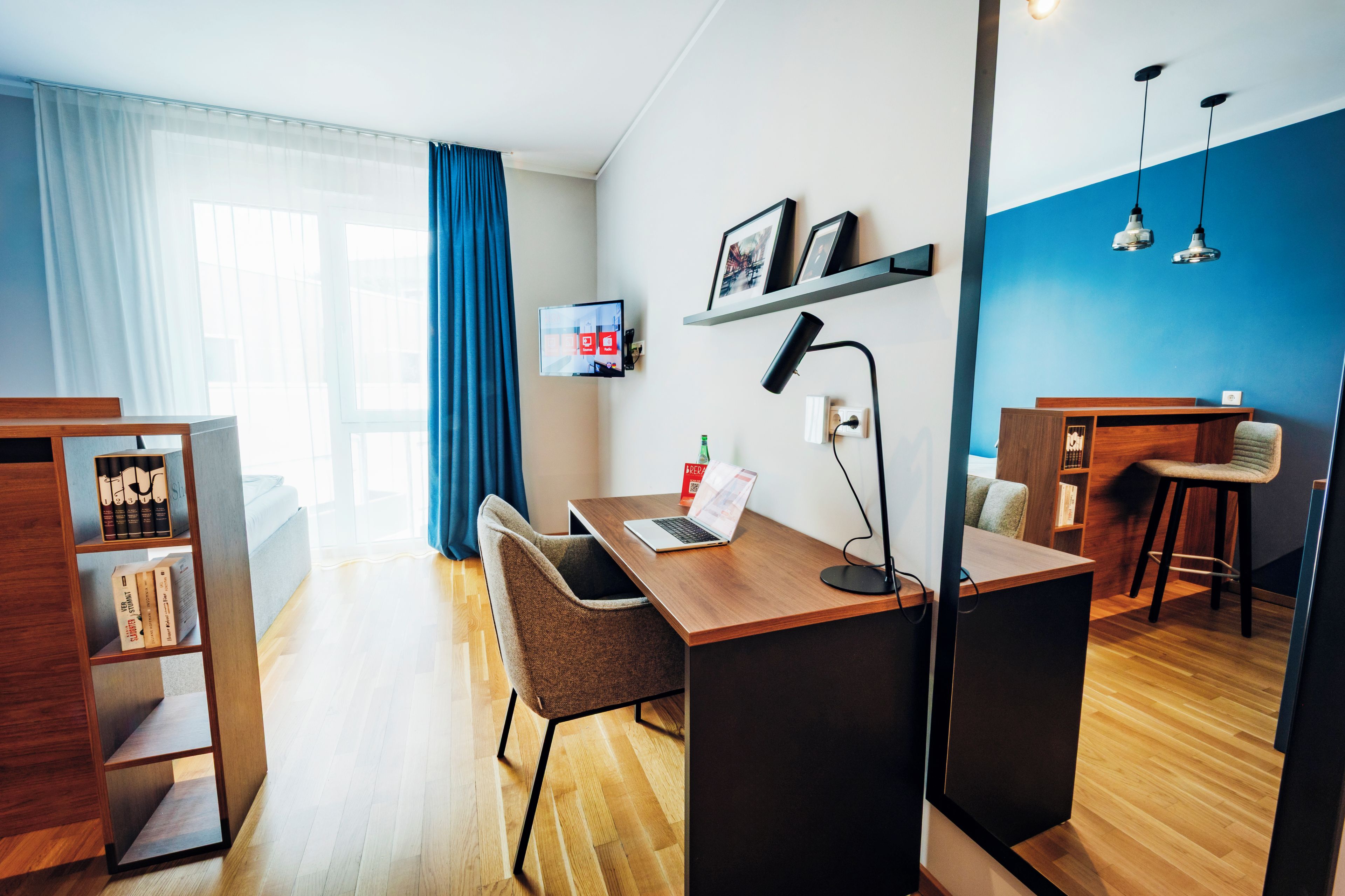 Ein modernes Wohnzimmer mit einem Holztisch, einem Laptop, einem Sessel, einem Bücherregal und einem an der Wand befestigten Fernseher.