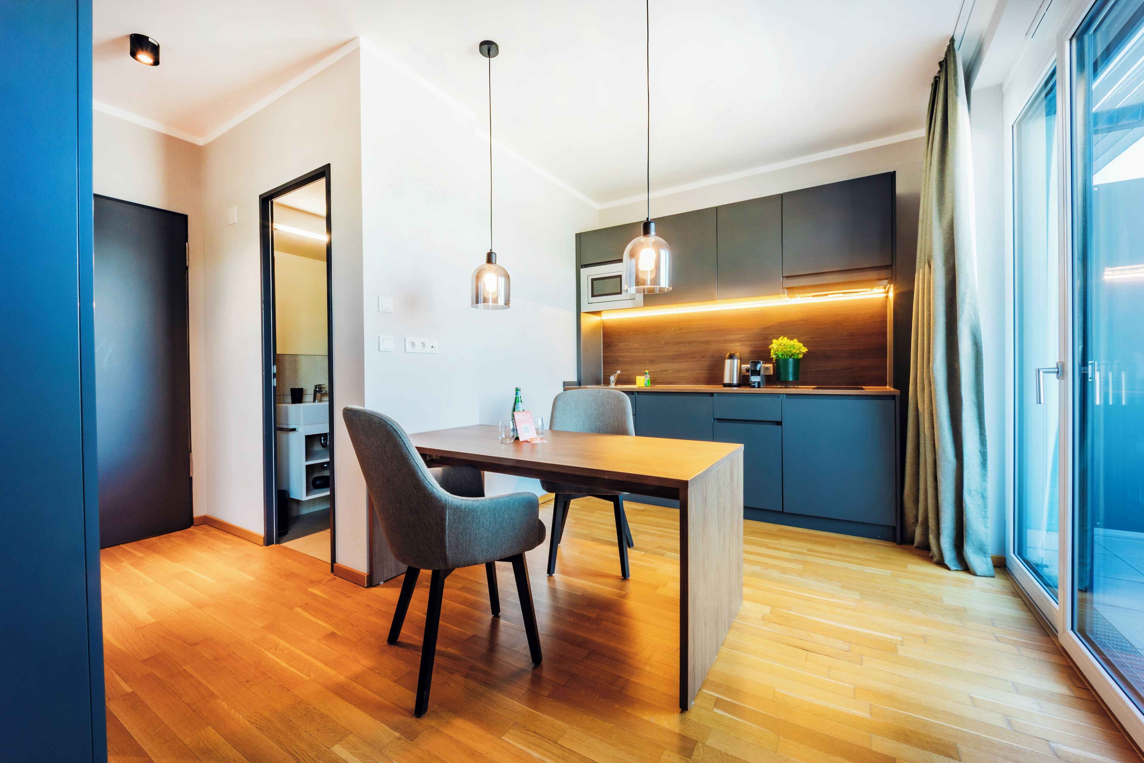 A modern kitchen with a dining area, wooden flooring, and a view through a large window.