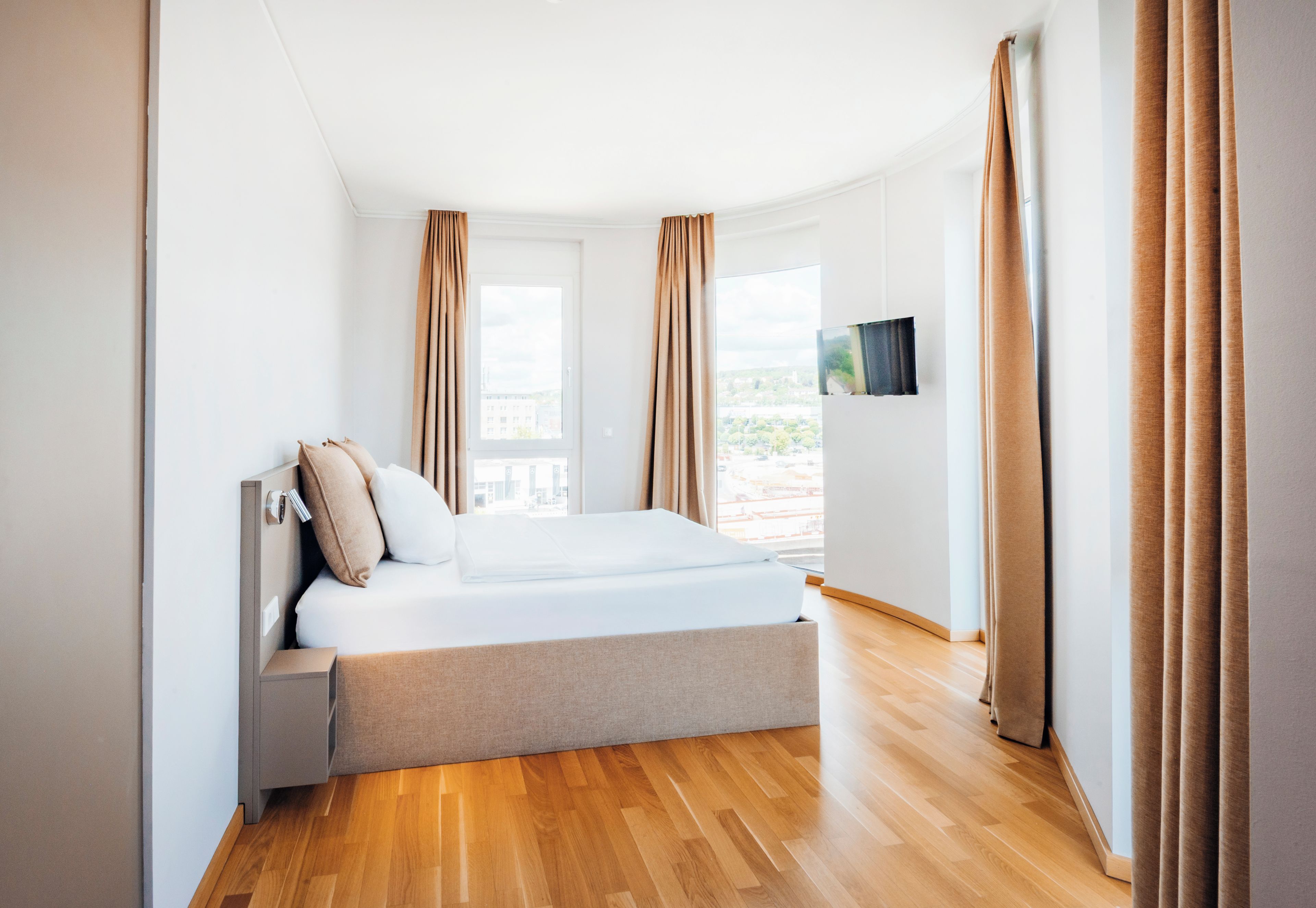 A modern bedroom with a bed, curtains, a TV, and a view of the city.
