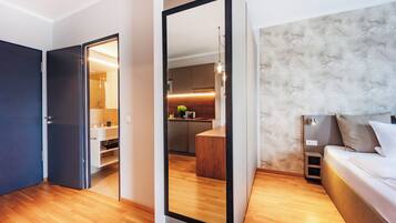 A modern hotel room with a wooden floor, a bed with a headboard, a large mirror, and a view into the adjacent kitchen area.