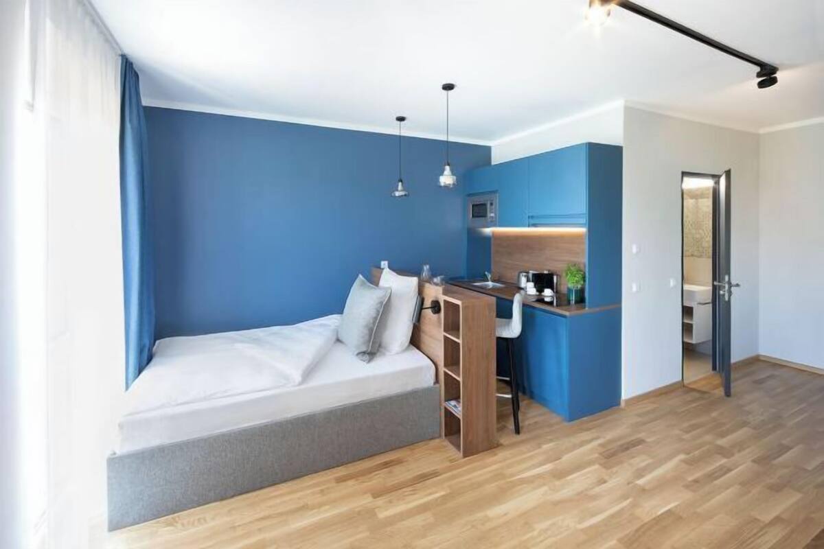A compact room with a bed, a kitchenette, and a door leading to another room.