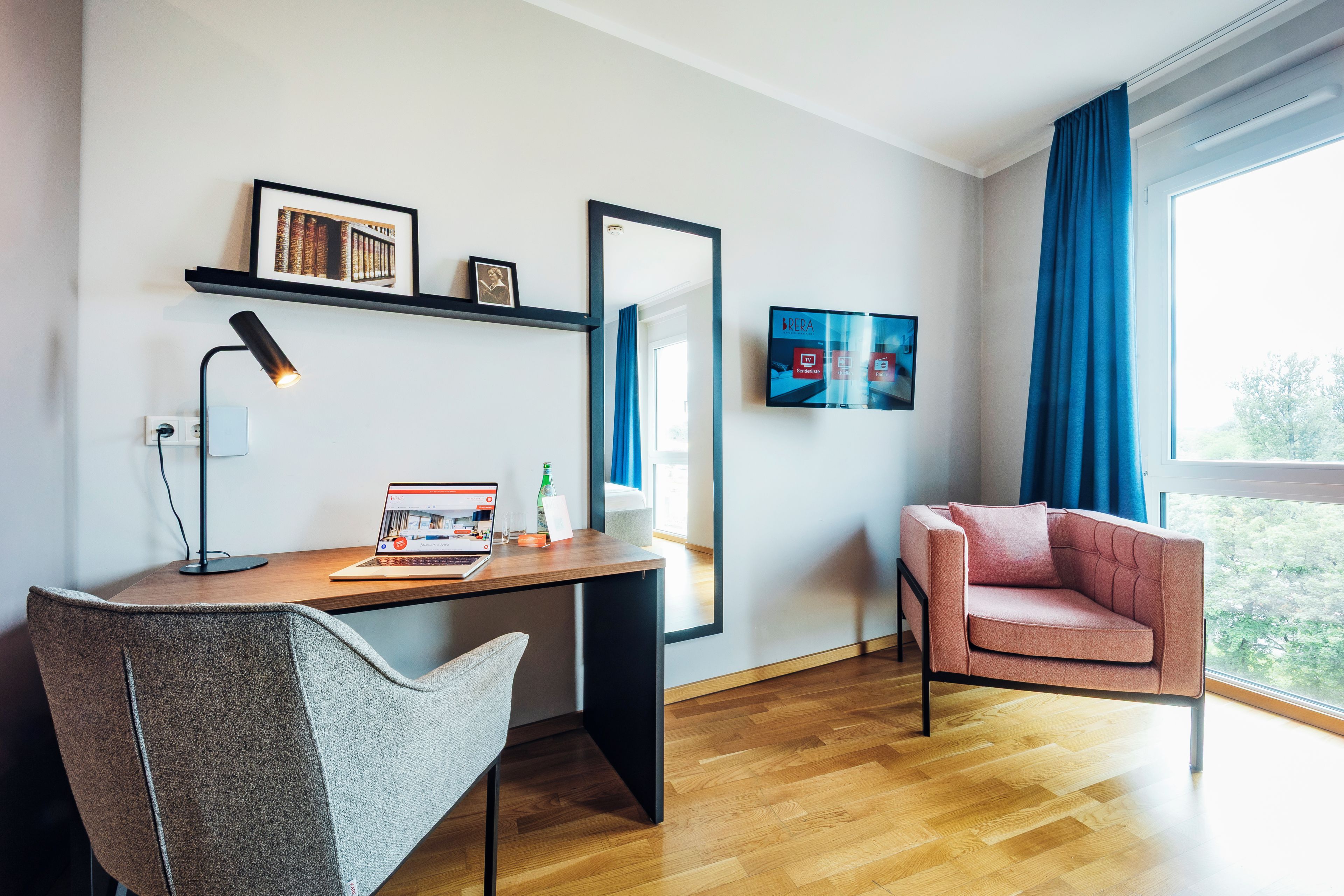 A modern hotel room with a wooden desk, a laptop, a chair, a large mirror, and a TV on the wall.