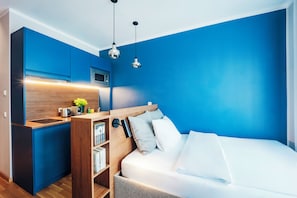 A modern bedroom with a built-in kitchenette, a bed with white bedding, and a blue accent wall. - Brera Serviced Apartments Ulm (Ulm)