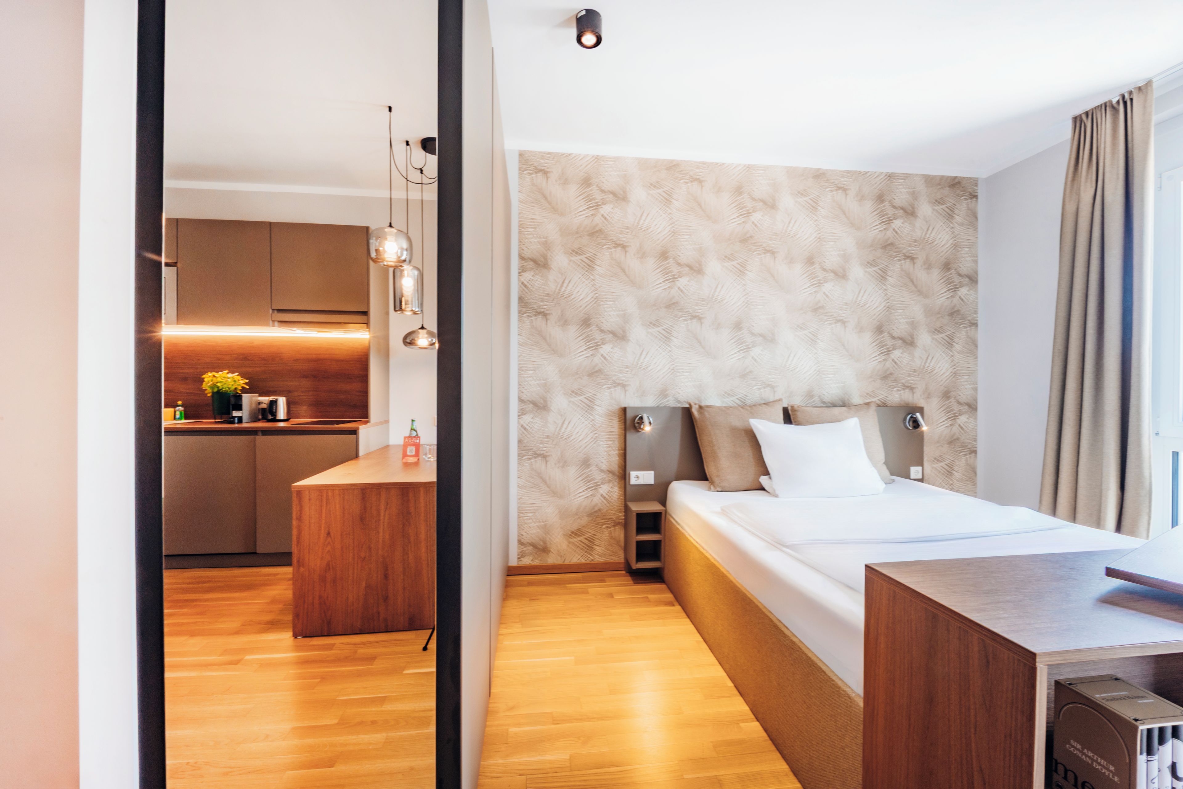A modern hotel room with a wooden floor, a bed with white linens, a desk, and a kitchenette in the background.