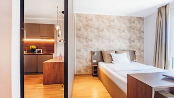 A modern hotel room with a wooden floor, a bed with white linens, a desk, and a kitchenette in the background.