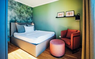 Una habitación de hotel moderna con una cama, un sillón rojo, un reposapiés redondo y una pared verde con cuadros.
