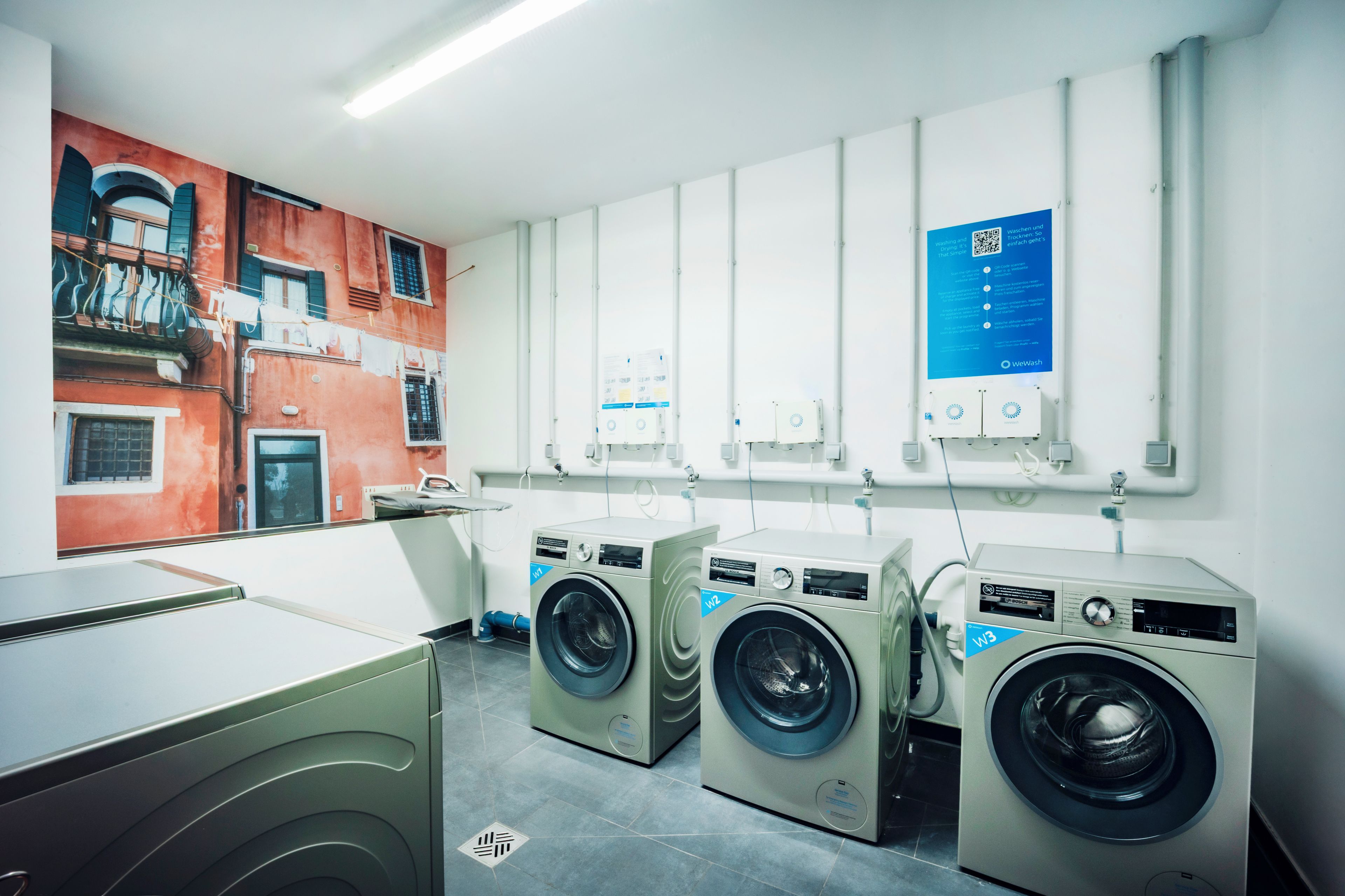 A laundry facility with three washing machines, a wall-mounted information board, and a view of a red building with balconies.