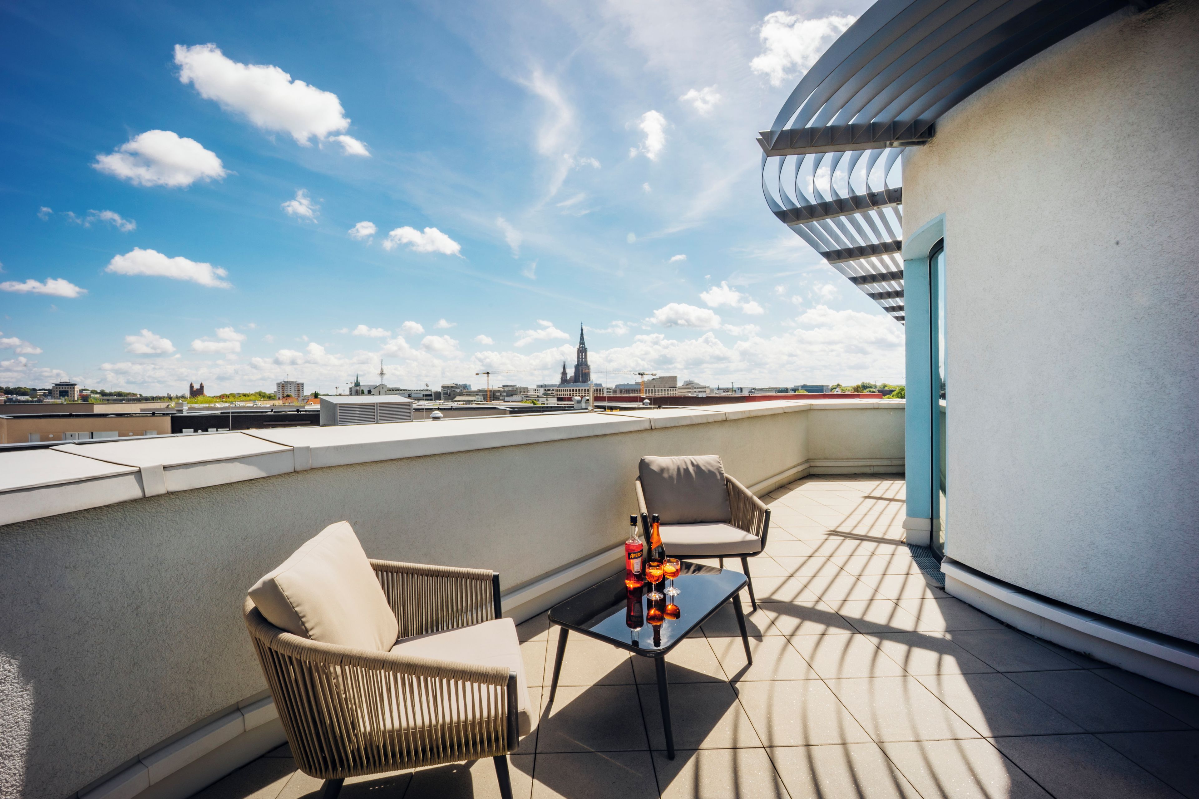 Une terrasse sur le toit, avec deux chaises et une table, offrant une vue dégagée sur les toits de la ville, et un bâtiment moderne doté d’un toit incurvé.