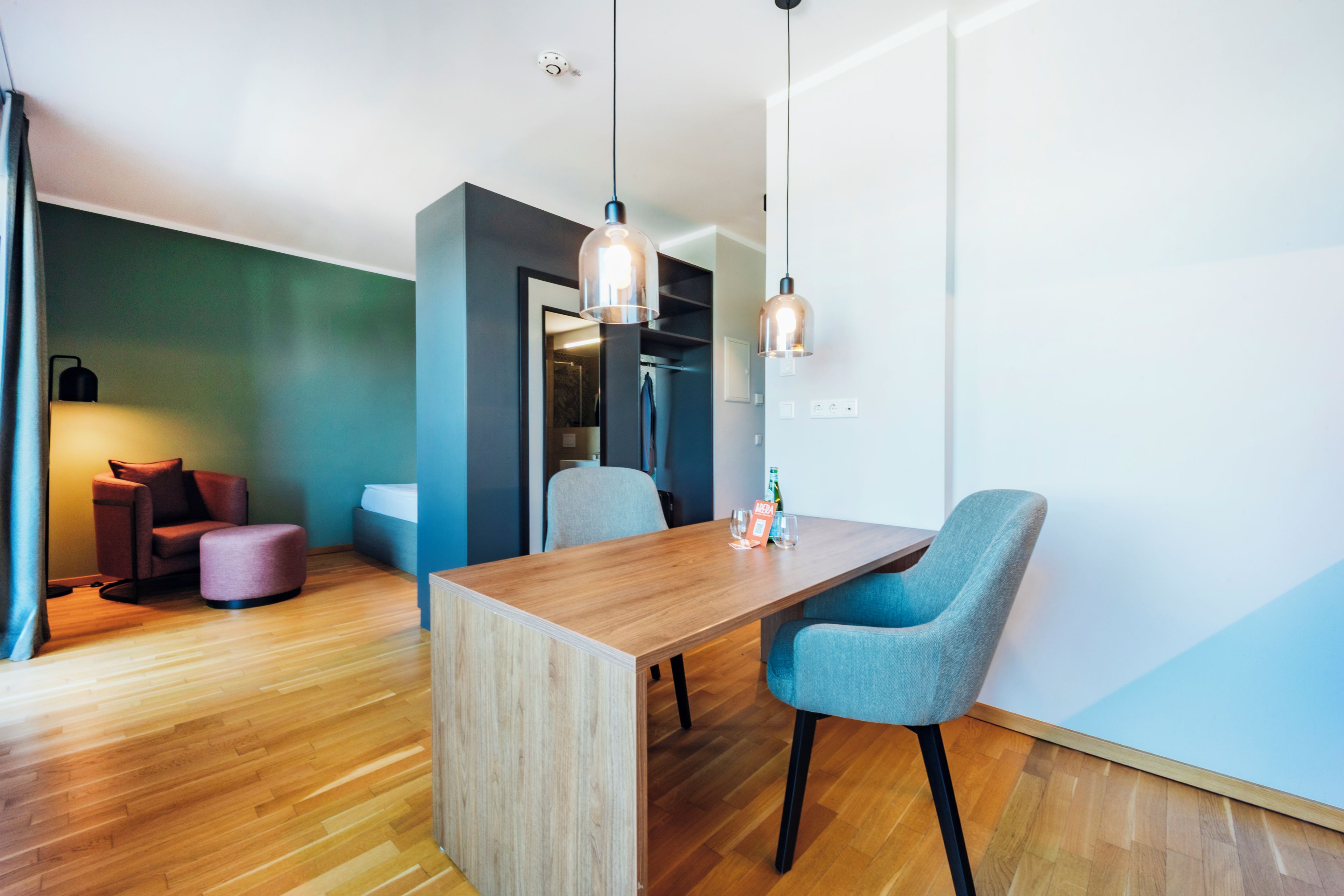A modern room with a wooden dining table, two blue chairs, a wooden floor, a green accent wall, and pendant lighting.
