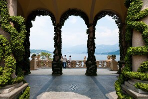 A covered walkway with arches, a view of a lake, and people walking. - ARIA Retreat & SPA - The Leading Hotels of the World (Porlezza)