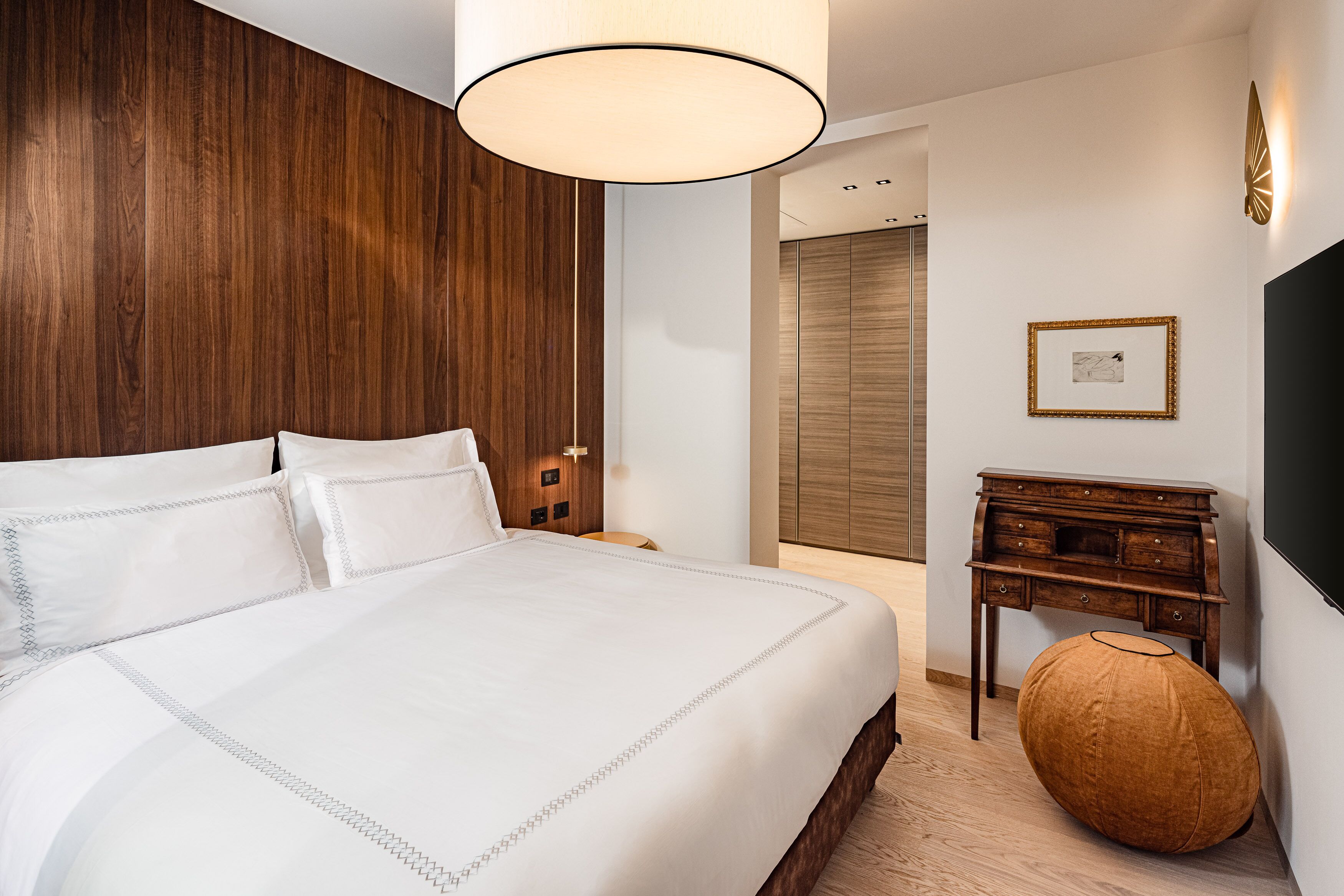 A hotel room with a large bed, wooden paneling on the wall, a small wooden dresser, and a round ottoman.