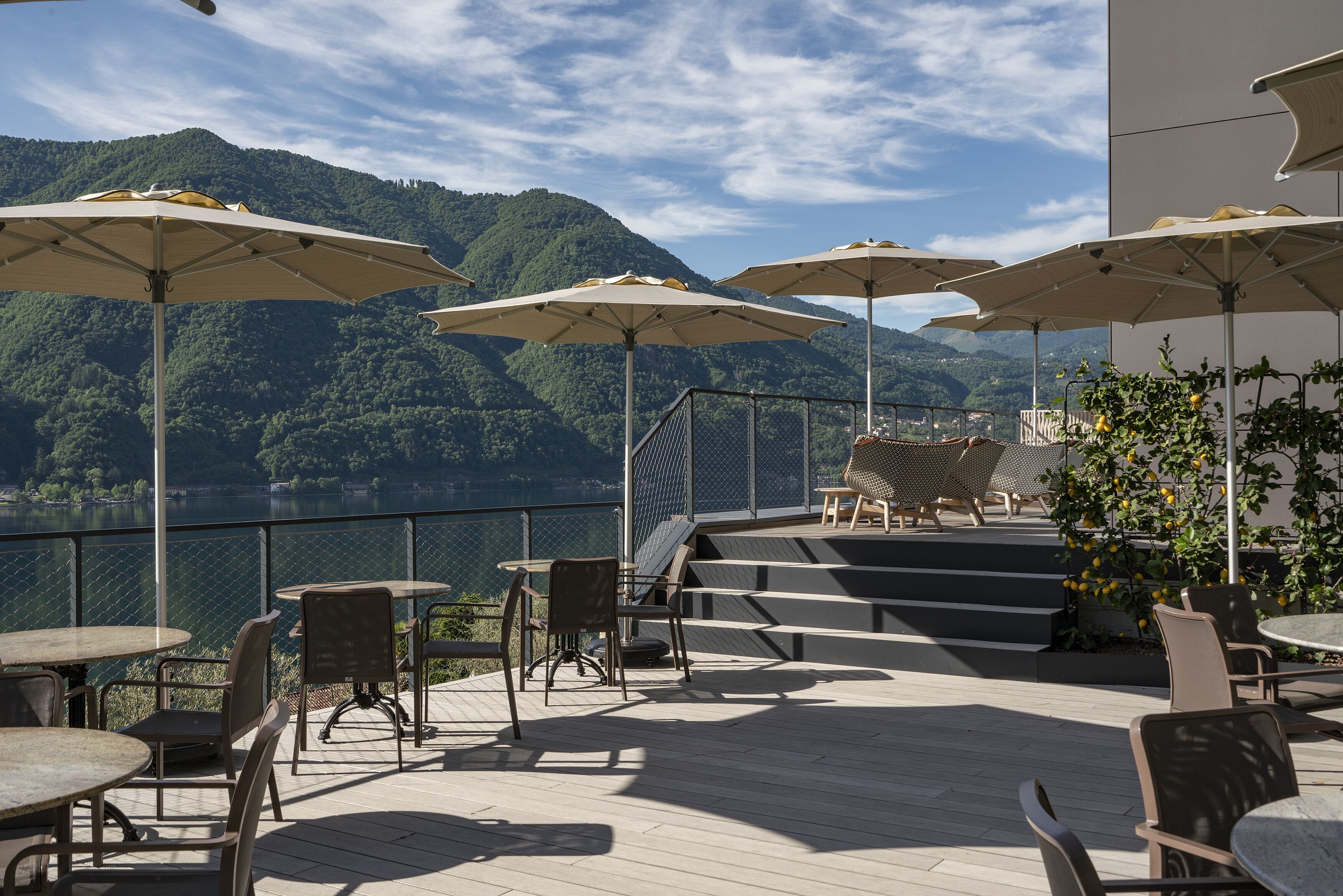 Une terrasse extérieure avec des tables et des chaises, des parasols, et une vue sur un lac et des montagnes.