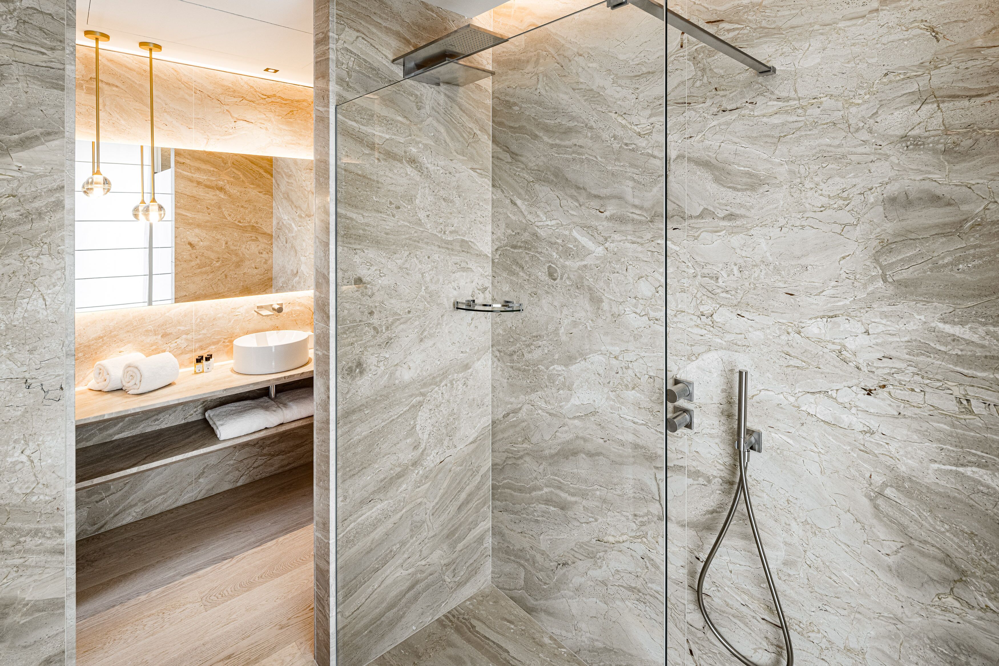 Un baño moderno con ducha envuelta en vidrio, paredes de mármol y un mueble de tocador de madera con toallas y artículos de aseo.