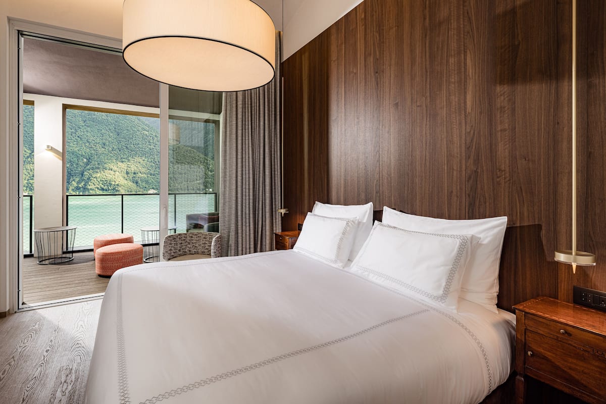 A hotel room with a large bed, wooden panel walls, a balcony with a view of a lake, and a modern chandelier.